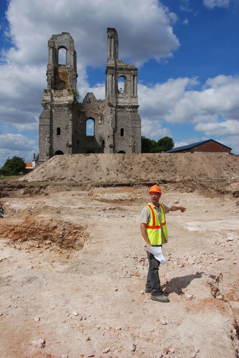 Vue du chantier de fouilles en 2010. Au premier plan un archéologue montre le cœur de l'abbatiale. En arrière plan, on peut appercevoir les 2 tours de l'abbaye du Mont Saint-Eloi.